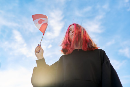 Student girl teenager with Canada flag in hand, blue sky with clouds background. Canada, education and youth, patriotism people conceptの写真素材