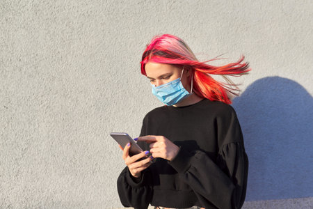 Hipster girl teenager with medical protective mask on face, with smartphone in hands. Gray outdoor...