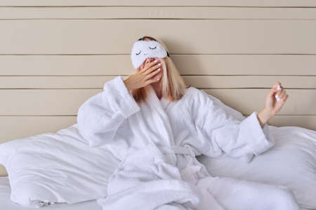 Young woman in white bed in bathrobe, eye mask, female yawns. Healthy lifestyle, sleep, rest, beauty conceptの写真素材