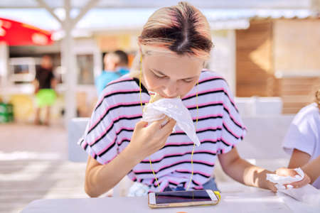 Fast food, summer, lifestyle, teenagers. Teen girl eating a sandwich, burger, looking at the smartphone screen in headphonesの写真素材