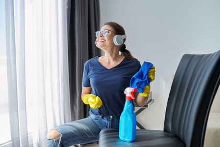 Woman doing house cleaning, smiling singing female in headphones and gloves with detergent washing furniture in room. Routine work in good mood, listening music, audio book, audio blogの写真素材
