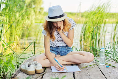 Creative teenager writes in notebook, girl sitting on wooden pier near lake in reeds, romantic dreamy female in her teens and experiences and interestsの写真素材