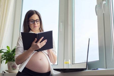 Young pregnant businesswoman working at home with laptop, female writing in business notebook,...
