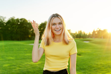 Gesturing young beautiful female teenager talking at camera, student 16, 17 years old giving interview looking at camera, green sunset lawn in park backgroundの写真素材
