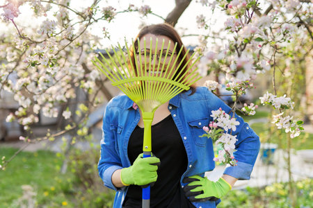 Spring gardening, portrait of mature smiling woman with rake looking at camera. Blooming trees in...