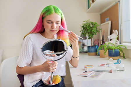 Trendy, fashionable teenager girl with dyed colored hair doing makeup looking in mirror with brush of decorative cosmetics sitting at table at homeの写真素材