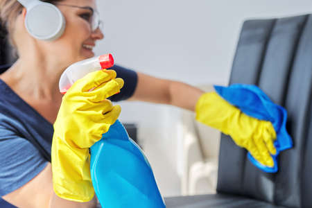 Home cleaning, woman in headphones and gloves with rag and spray with detergent washing leather chair. Housework, housekeeping, household, cleanliness conceptの写真素材