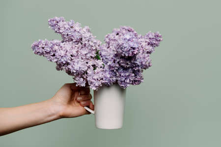 Lilac flowers in white cup in the hands of woman on green background. Spring, beauty, perfumery, cosmetology, holiday, gift, decoration conceptの写真素材