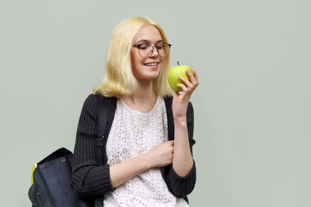 Teenager girl student with backpack, with green apple in her hand on light background. Teens,...