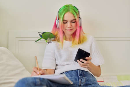 Bright beautiful owner of parrot teenager student with pet on her shoulder. Trendy dyed hair girl with headphones writing in a school notebook, using smartphone to studyの写真素材