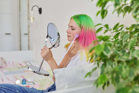 Fashionable teenage model with colored dyed hair doing makeup using mirror makeup brush, sitting at home in bed. Female with pink green yellow hair, fashion trends hairstyles beautyの写真素材