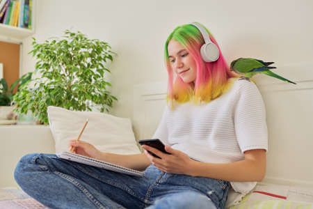Bright beautiful owner of parrot teenager student with pet on her shoulder. Trendy dyed hair girl with headphones writing in a school notebook, using smartphone to studyの写真素材