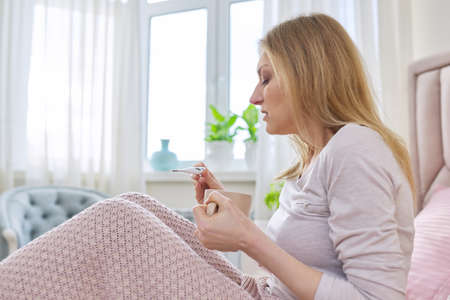 Middle aged woman with thermometer sitting at home in bed under blanketの写真素材