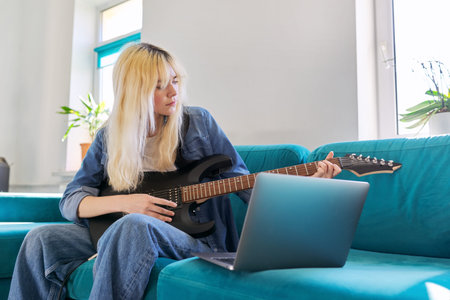 Hipster teenager girl playing electric guitar looking at laptop monitor