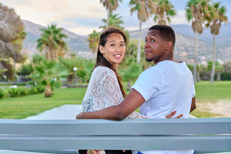 Beautiful young multi-ethnic couple sitting together on a bench in a tropical parkの写真素材