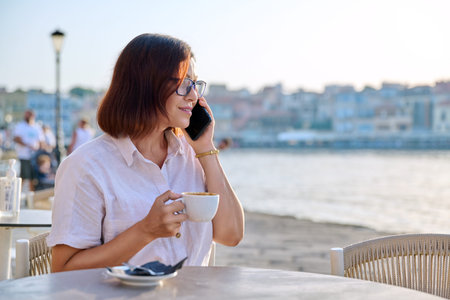 Mature woman in outdoor restaurant on seafront with cup of coffee talking on a smartphone.の写真素材