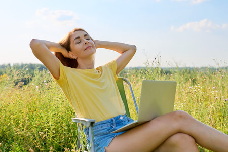 Adult woman sitting on an folding camping chair using a laptop, in a meadow with wild herbs