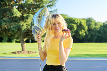 Portrait of beautiful teenage blonde with cake donut and silver balloon in park