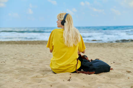 Teenage female in headphones with backpack sitting on beach, back view, copy spaceの写真素材