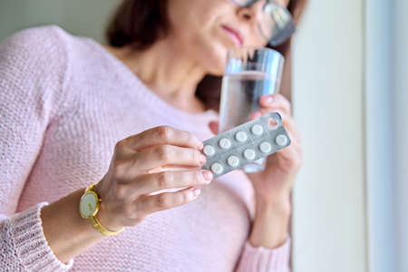 Mature sad woman with blister of pills and glass of water at home.の写真素材