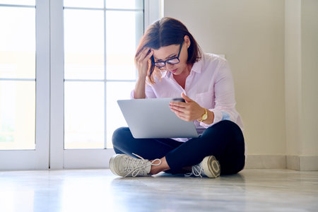 Serious middle aged woman sitting on floor using laptop