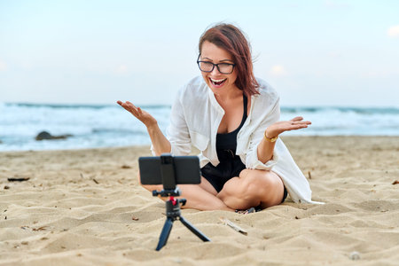 Middle aged woman sitting on beach with smartphone using video callの写真素材