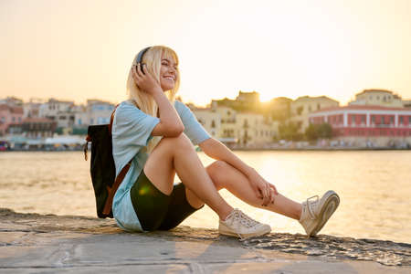 Happy teenage female tourist sitting on sea promenade of an old city in sunsetの写真素材