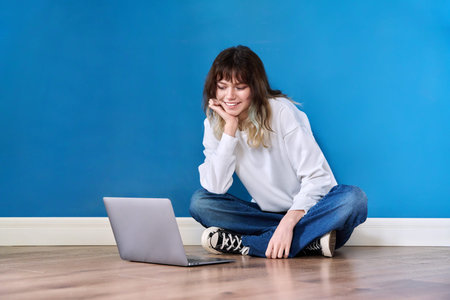 Beautiful teenage female sitting on the floor with laptop on blue background