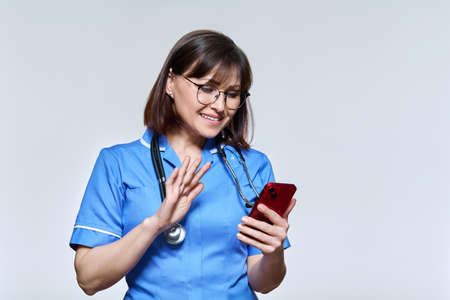 Friendly nurse waving hand at smartphone screen, on light studio backgroundの写真素材