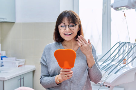 Happy woman dentist patient with mirror in hands looking at her teethの写真素材