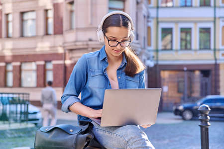 Teenage student girl in headphones using laptop outdoor, urban backgroundの写真素材