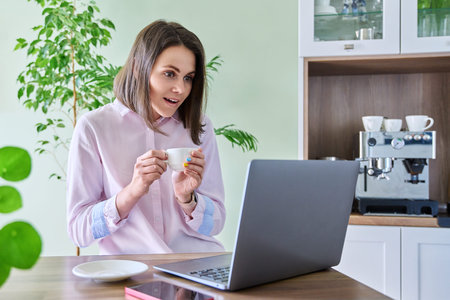 Joyful surprised woman looking at laptop screen, sitting at home in kitchen