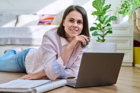 Attractive young woman looking at camera lying on floor at home with laptop