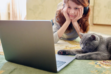 Preteen girl in headphones and gray british cat looking at laptop together