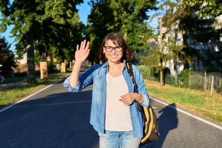 Happy woman with backpack waving hand looking at camera, on roadの写真素材