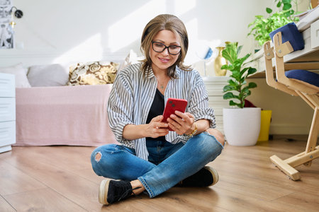 Middle aged woman sitting on floor at home with smartphone
