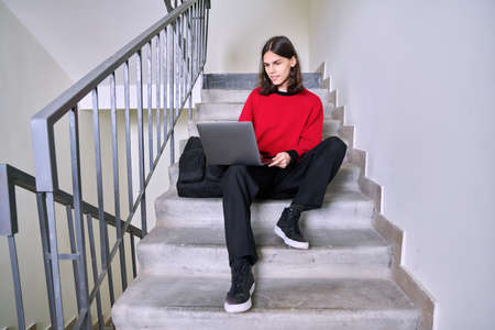 Portrait of student guy on the steps inside building using laptopの写真素材