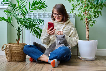 Woman at home using smartphone, warming with cat near heating radiatorの写真素材