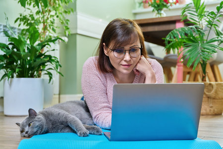Middle-aged woman lying with cat on exercise mat, looking at laptop