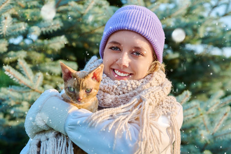 Young female in snowy weather with cat looking at camera, outdoor near Christmas tree