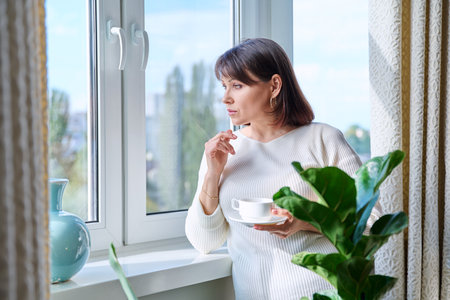 Middle aged woman at home near the window with a cup of coffeeの写真素材