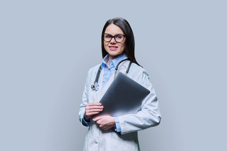 Young friendly female doctor with laptop on light gray backgroundの写真素材