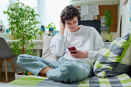 Young male in headphones looking in smartphone while sitting on couch at homeの写真素材