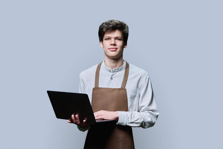 Young male in an apron with laptop, on gray studio backgroundの写真素材