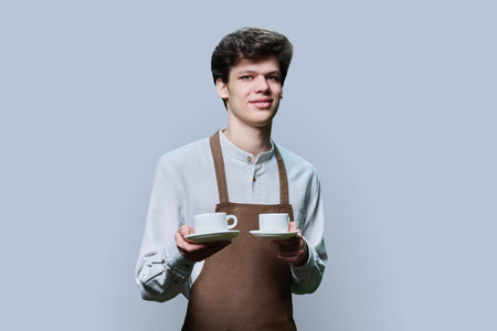 Young male waiter in apron posing on gray background with two cups of coffeeの写真素材