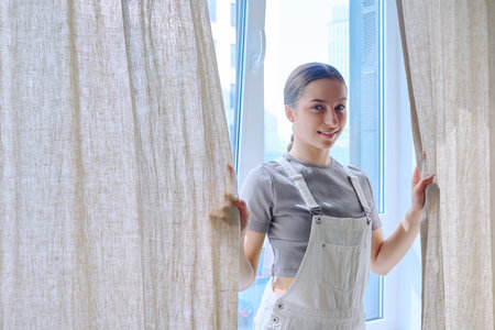 Smiling teenage girl looking at camera standing near window with curtainsの写真素材