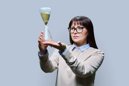 Mature woman holding an hourglass in her hands, on gray backgroundの写真素材