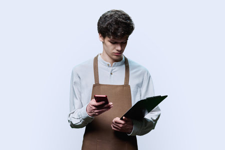 Portrait of young guy in apron with clipboard, smartphone on white backgroundの写真素材