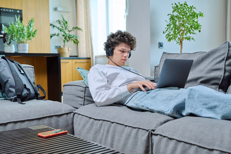 Young guy lying on sofa in living room, looking at laptop screen in headphonesの写真素材