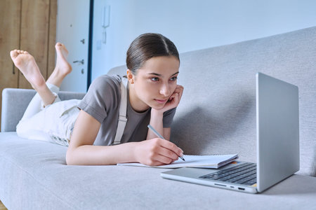 Teen girl at home on couch using laptop, writing in a notebook